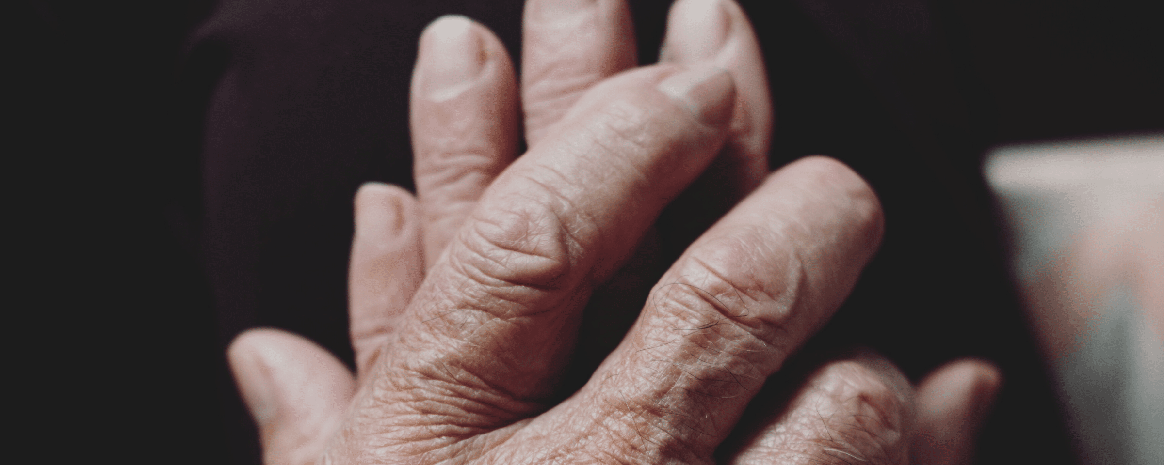 A imagem mostra o detalhe das mãos entrelaçadas de uma pessoa idosa, com destaque para as rugas e texturas da pele, indicando idade avançada. O fundo é escuro, focando toda a atenção para as mãos. No topo da imagem, há texto que parece ser o anúncio de um evento, lê-se: "EVENTO INTERNACIONAL Simpósio aborda Saúde Integrativa para Saúde Mental do Idoso". No canto inferior esquerdo, há um pequeno ícone que parece ser uma árvore estilizada dentro de um quadrado, possivelmente um logotipo relacionado ao evento ou à organização responsável. A composição da imagem e o texto sugerem um foco na importância da saúde mental integrativa para a população idosa, promovida por meio de um evento internacional dedicado ao tema.
