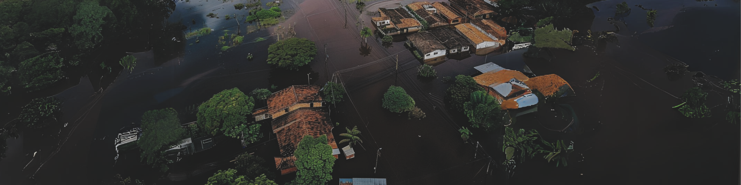 A imagem aérea mostra uma extensa área residencial inundada. As águas marrons submergiram ruas e cercaram casas com telhados de barro, evidenciando o impacto severo das inundações. Algumas árvores emergem das águas, que refletem parcialmente as casas e a vegetação.